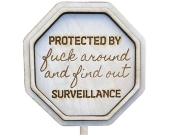 May include: Wooden sign with an octagonal shape and a stick. The sign has the words "PROTECTED BY fuck around and find out SURVEILLANCE" engraved in a dark brown color.