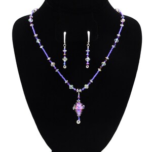 May include: A purple and silver crystal necklace with a dangling iridescent glass pendant and matching earrings. The necklace is on a black velvet display stand.