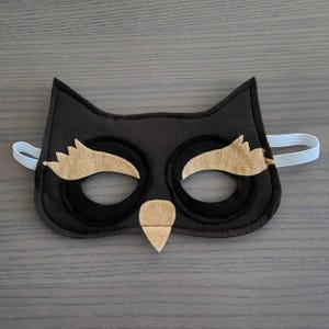 May include: A black felt owl mask with tan detailing and a white elastic strap. The mask has large eye holes, tan eyebrows, and a beak. Ideal for fancy dress or imaginative play, this is a handmade item.