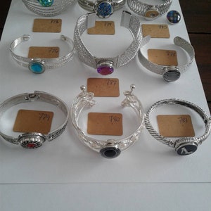 Puede incluir: Una colección de pulseras plateadas con diversos diseños, incluyendo estilos trenzados, de eslabones y texturizados. Algunas pulseras presentan acentos de piedras preciosas coloridas. Cada pulsera tiene una pequeña etiqueta numerada.