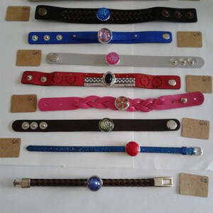 May include: A collection of ten leather bracelets with different coloured and patterned bands and decorative snap closures. The bracelets are displayed on a white surface with brown paper tags.