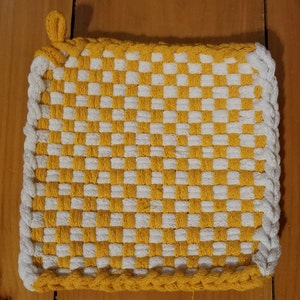May include: A square, woven potholder with a yellow and white checkerboard pattern. The edges are trimmed with a white, braided border and a yellow loop for hanging. The potholder is designed for kitchen use.