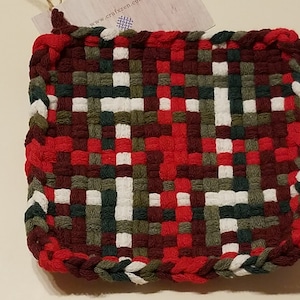 May include: A square, woven hot pad in a patchwork design. The pad features a pattern of red, white, green, and dark red squares. The edges are finished with a braided border in red, white, and green. A tag is attached to the top.