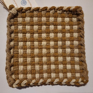May include: A square woven potholder featuring a checkered pattern in alternating shades of mustard yellow and cream. The edges are finished with a thick, braided border in a solid tan color, and a small paper tag is attached to the top corner.
