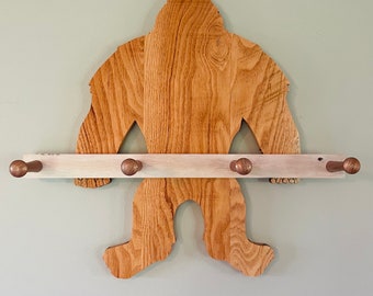 May include: Wooden wall-mounted coat rack shaped like a Bigfoot with four hooks. The rack is made of natural wood with a light brown stain.