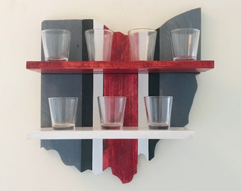 May include: A wooden shelf shaped like the state of Ohio, painted in red, white and grey stripes. The shelf has two tiers and holds six clear glass shot glasses.