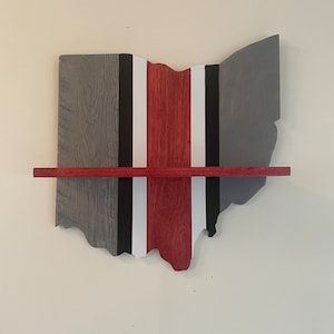 Puede incluir: Estante de madera con forma de Ohio, pintado en gris, negro, blanco y rojo. Un estante horizontal se extiende por el centro. El estante está montado en una pared de color crema.