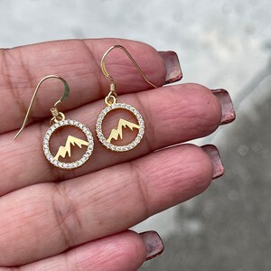 May include: Gold earrings with a circle design featuring a mountain silhouette and clear cubic zirconia stones.