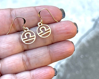Könnte beinhalten: Ohrringe in Goldton mit dem Sternzeichen Waage. Die Ohrringe sind klein und zart, mit einem runden Design, das das Waage-Symbol zeigt.