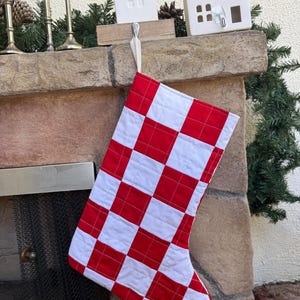 May include: A quilted Christmas stocking featuring a vibrant red and white checkered pattern. The stocking is constructed from square fabric patches stitched together with a visible grid pattern and includes a simple fabric loop for hanging.