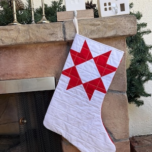 May include: A quilted Christmas stocking featuring a white fabric base with a prominent red eight-pointed star pattern on the front. The stocking is constructed with horizontal stitching lines and includes a small white fabric loop for hanging.