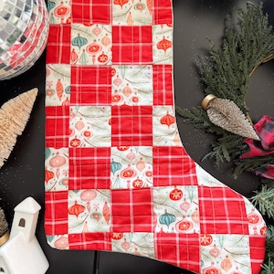 May include: A quilted Christmas stocking with a patchwork design. The stocking features red plaid squares alternating with squares printed with Christmas ornaments in red, pink, and teal. A white loop is attached at the top for hanging.