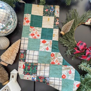 May include: A quilted Christmas stocking with a patchwork design. The stocking features squares of green, cream, and patterned fabrics with holiday motifs like ornaments and ballerinas. The stocking is surrounded by Christmas decorations.