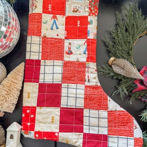 May include: A quilted Christmas stocking with a patchwork design in red and cream. The squares feature festive patterns like trees, stars, and figures. The stocking is decorated with a variety of holiday-themed images and patterns.