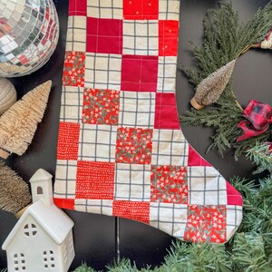 May include: A quilted Christmas stocking with a patchwork design. The stocking features red and white squares, some with a floral pattern, and is trimmed with red fabric. The background includes Christmas decorations.