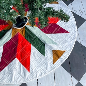 May include: A white quilted Christmas tree skirt with a geometric pattern in red, green, gold, and burgundy. The skirt is decorated with a starburst design and has a white trim.