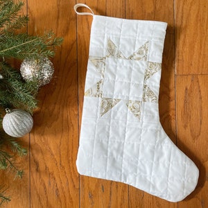 May include: White quilted Christmas stocking with a gold star design. The star is made of gold fabric with a leaf pattern. A loop is attached for hanging. Christmas decor.