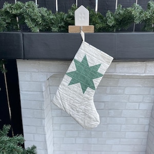 May include: A white quilted Christmas stocking with a green star design hanging from a white ceramic house-shaped stocking holder.