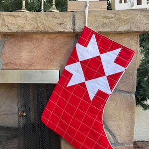 May include: A quilted Christmas stocking featuring a vibrant red fabric base with a white grid pattern. The upper section displays a large, white, eight-pointed star design centered against the red background. It includes a white fabric hanging loop.