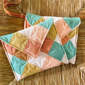 May include: A quilted clutch with a geometric star pattern in shades of peach, yellow, teal, and white. The clutch has a flap closure and a brown strap with star details. The bag is resting on a woven surface.
