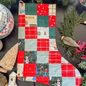 May include: A quilted Christmas stocking with a patchwork design. The stocking features squares of red, green, and light blue fabric, some with star patterns, plaid, and holiday-themed illustrations. The top has a loop for hanging.