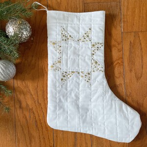 May include: White quilted Christmas stocking with a gold star design. The star is made of fabric with a gold glitter pattern.