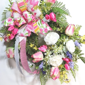May include: A spring wreath featuring pink tulips, white ranunculus, and various greenery. A large bow with pink, white, and yellow striped ribbon accents the top. Pink and white ribbons hang down, adding to the wreath's decorative appeal.
