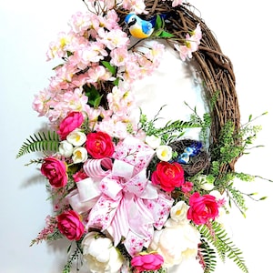 May include: A spring wreath with pink flowers, white flowers, green leaves, and a pink and white ribbon bow. Two blue birds are perched on a branch and a bird's nest with two blue birds is on the wreath.
