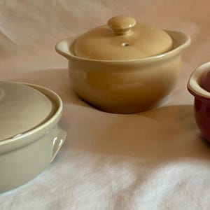 May include: Three ceramic lidded bowls in neutral tones. One is taupe, one is tan, and the other is a deep burgundy. Each bowl has a matching lid with a small knob and a vent hole. The bowls are oval or round, with a slightly flared rim.