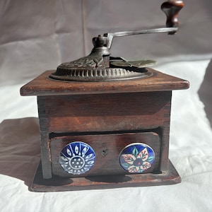 May include: A vintage wooden coffee grinder with a metal crank and decorative floral knobs on the drawer. The grinder is made of dark wood and has a classic design, perfect for kitchen decor.