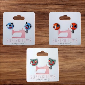 May include: Three pairs of stud earrings on white cards. The earrings feature cartoon faces in blue, orange, and green. Each card has a pink sewing machine graphic and the text "SUPERKITTY'S sewing & crafts".