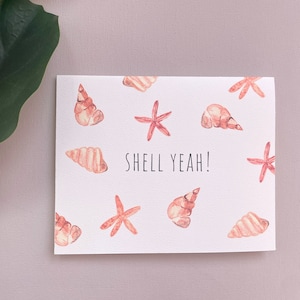May include: A white card with a watercolor illustration of seashells and starfish. The text "Shell Yeah!" is written in black on the card.