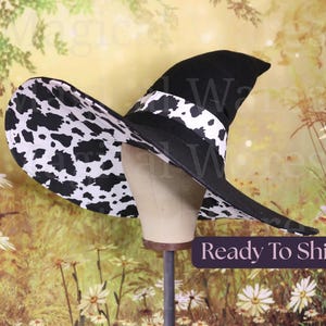 May include: A pointed witch hat featuring a solid black exterior and a wide brim lined with a black and white cow print pattern. A matching cow print band encircles the base of the crown, contrasting against the black fabric of the hat.