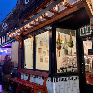 Looking inside the windows of a cafe at night. Inside the cafe are three macrame plant hangers and macrame wall hangings on a white wall.