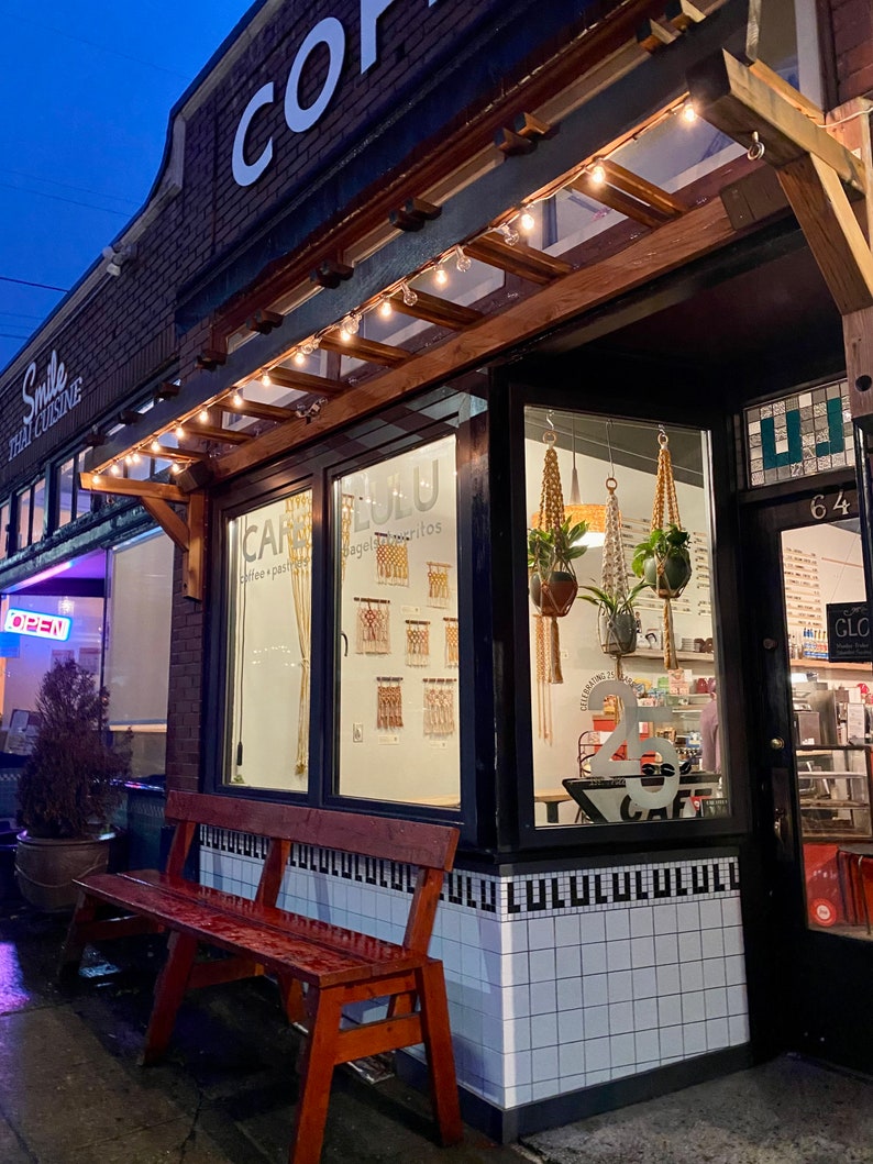 Looking inside the windows of a cafe at night. Inside the cafe are three macrame plant hangers and macrame wall hangings on a white wall.