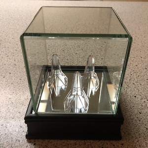 May include: Three clear glass crystal figurines displayed in a glass cube with a mirrored base and a black wooden frame.