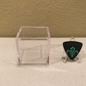 May include: A clear plastic display case with a black guitar pick featuring a green fleur-de-lis design and the text "CROWBAR".