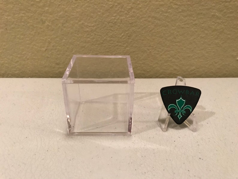 May include: A clear plastic display case with a black guitar pick featuring a green fleur-de-lis design and the text "CROWBAR".
