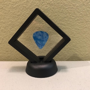 May include: A blue guitar pick displayed in a black plastic display case with a clear plastic cover. The case has a black base.