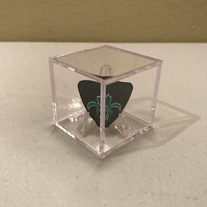 May include: A clear plastic cube display case with a black and green guitar pick inside. The pick has a fleur-de-lis design.