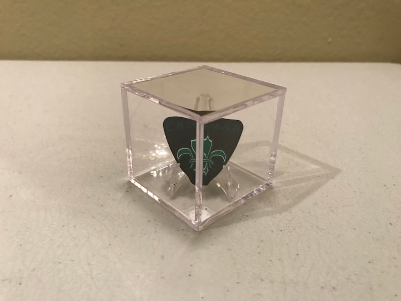 May include: A clear plastic cube display case with a black and green guitar pick inside. The pick has a fleur-de-lis design.