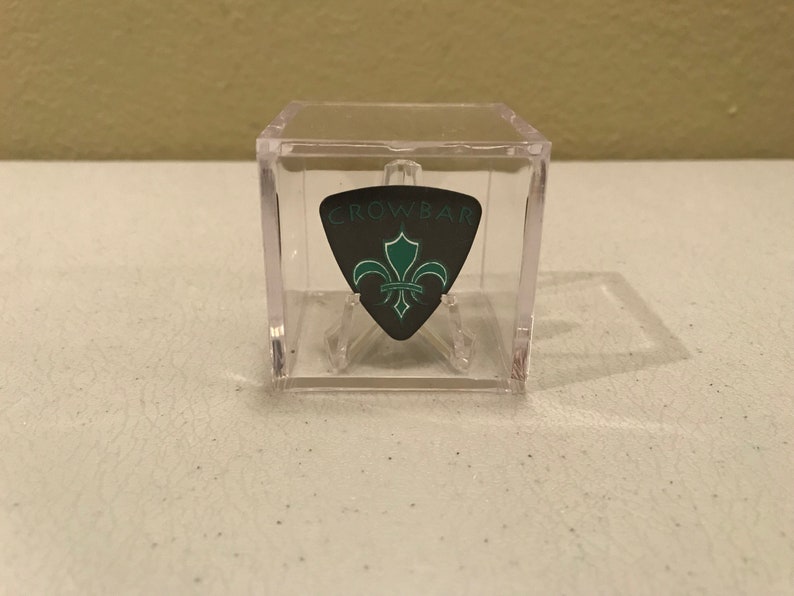 May include: A black guitar pick with a green fleur-de-lis design and the word "CROWBAR" in white lettering. The pick is displayed in a clear plastic cube.