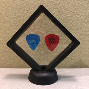 May include: A black diamond-shaped display case with a clear acrylic insert, showcasing two guitar picks. One pick is blue and the other is red with the text "Dunlop" printed on it.