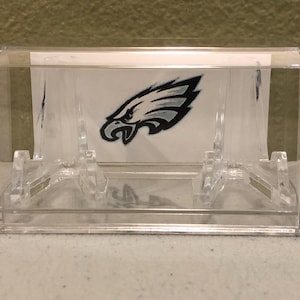 May include: A clear plastic display case with a white background and a black and white Philadelphia Eagles logo. The case has two clear plastic stands to hold items.