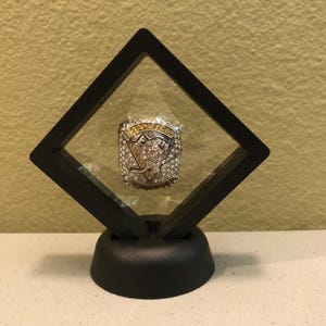 May include: A black diamond-shaped display case with a clear glass top, showcasing a silver and gold championship ring with a team logo and gemstones. The case sits on a black base.
