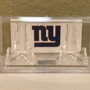 May include: A clear rectangular acrylic display case containing two transparent plastic stands. Inside the case, a white card features the dark blue lowercase letters ny in a bold font, representing the New York Giants sports team logo.