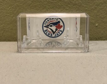 May include: A clear plastic display case with a white card inside featuring the Toronto Blue Jays logo. The logo is a blue circle with a white bird and baseball, and the words "Toronto Blue Jays" in white.