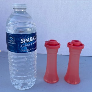 May include: Two pink plastic bottle stoppers with a clear plastic bottle of water in the background. The bottle of water is labeled "Sparkle Purified Water" and has a label that says "Please Recycle Non-Carbonated".
