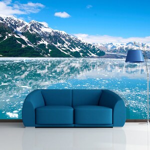 May include: A blue sofa and a blue lamp stand in front of a wall mural depicting a mountain range with snow-capped peaks reflected in a body of water. The sky is a bright blue with a few clouds.