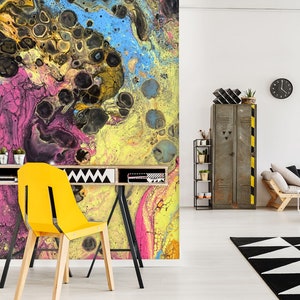 Op de afbeelding: Abstracte wandmuur met gele, roze, blauwe en zwarte verfswirls. De muurschildering bevindt zich in een moderne woonkamer met een gele stoel, een bureau en een metalen locker.